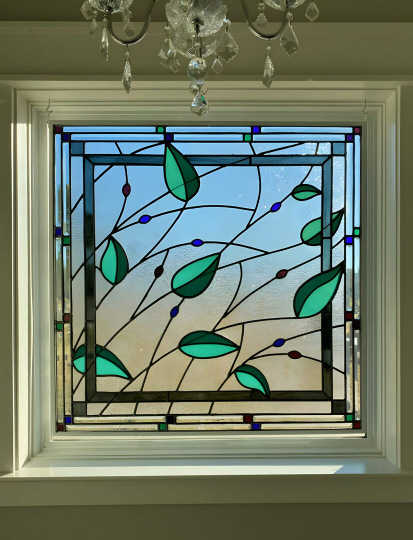 Stained glass window with leaf design set in a white frame against a clear sky.