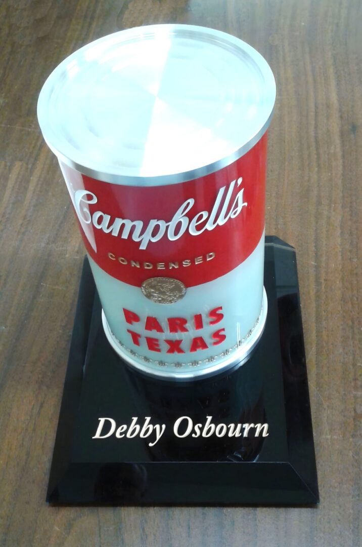 A campbell's condensed soup can-themed award with a nameplate reading "debby osbourn" displayed on a black base with "paris texas" inscribed.