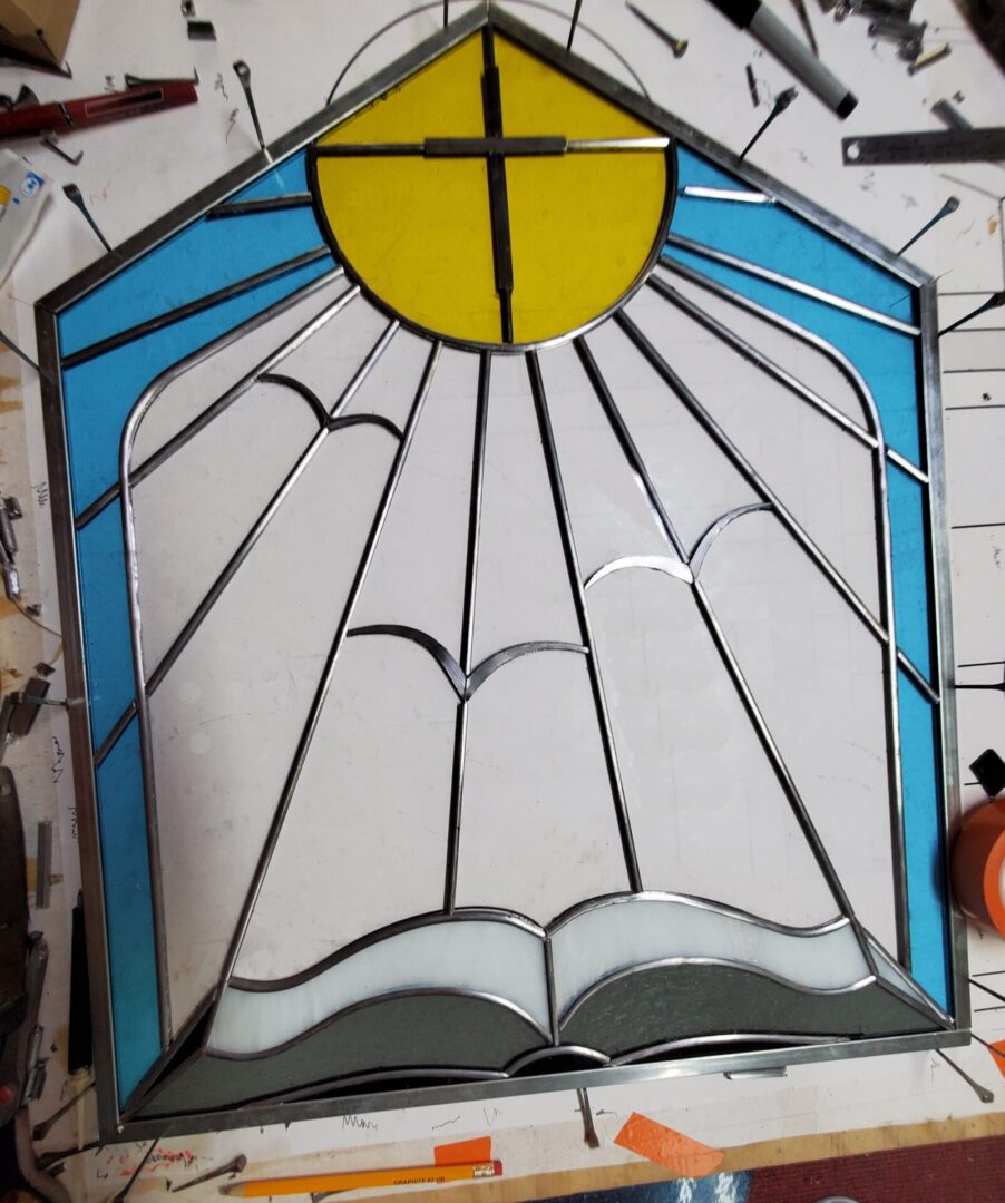 Stained glass artwork of a sunrise or sunset with a cross in the center, placed on a work table amidst craft tools and materials.