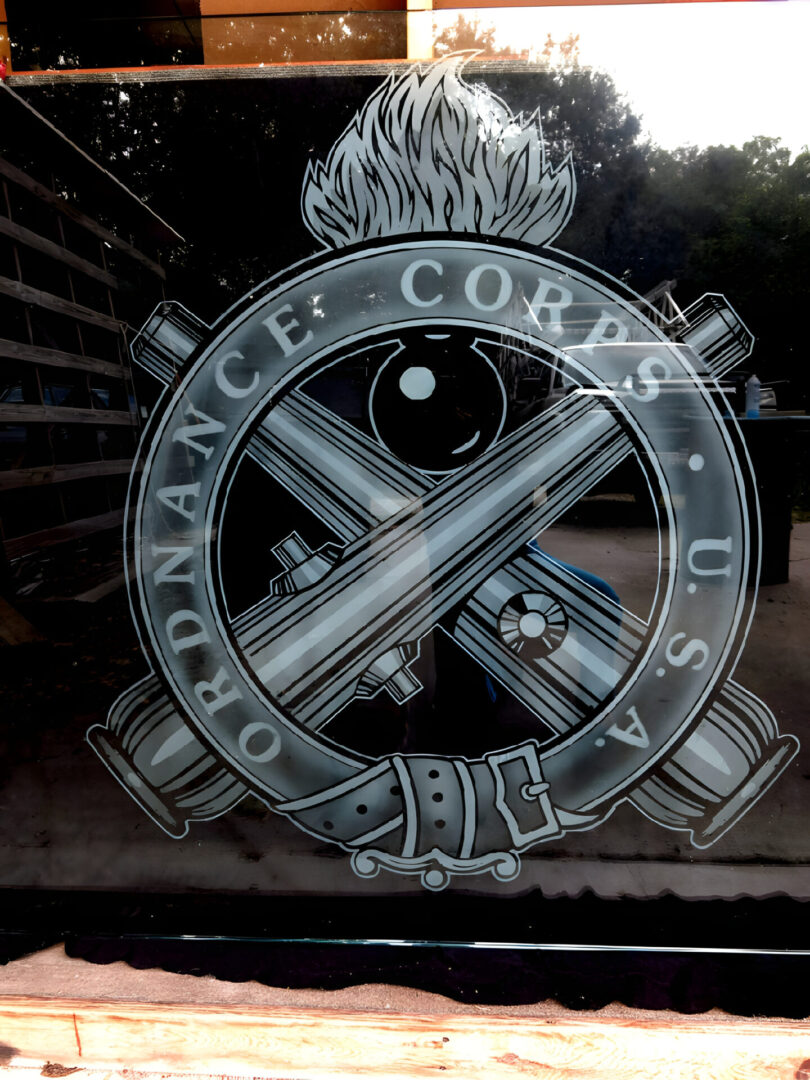 Emblem of the u.s. army ordnance corps etched on a glass surface with a reflection of trees in the background.