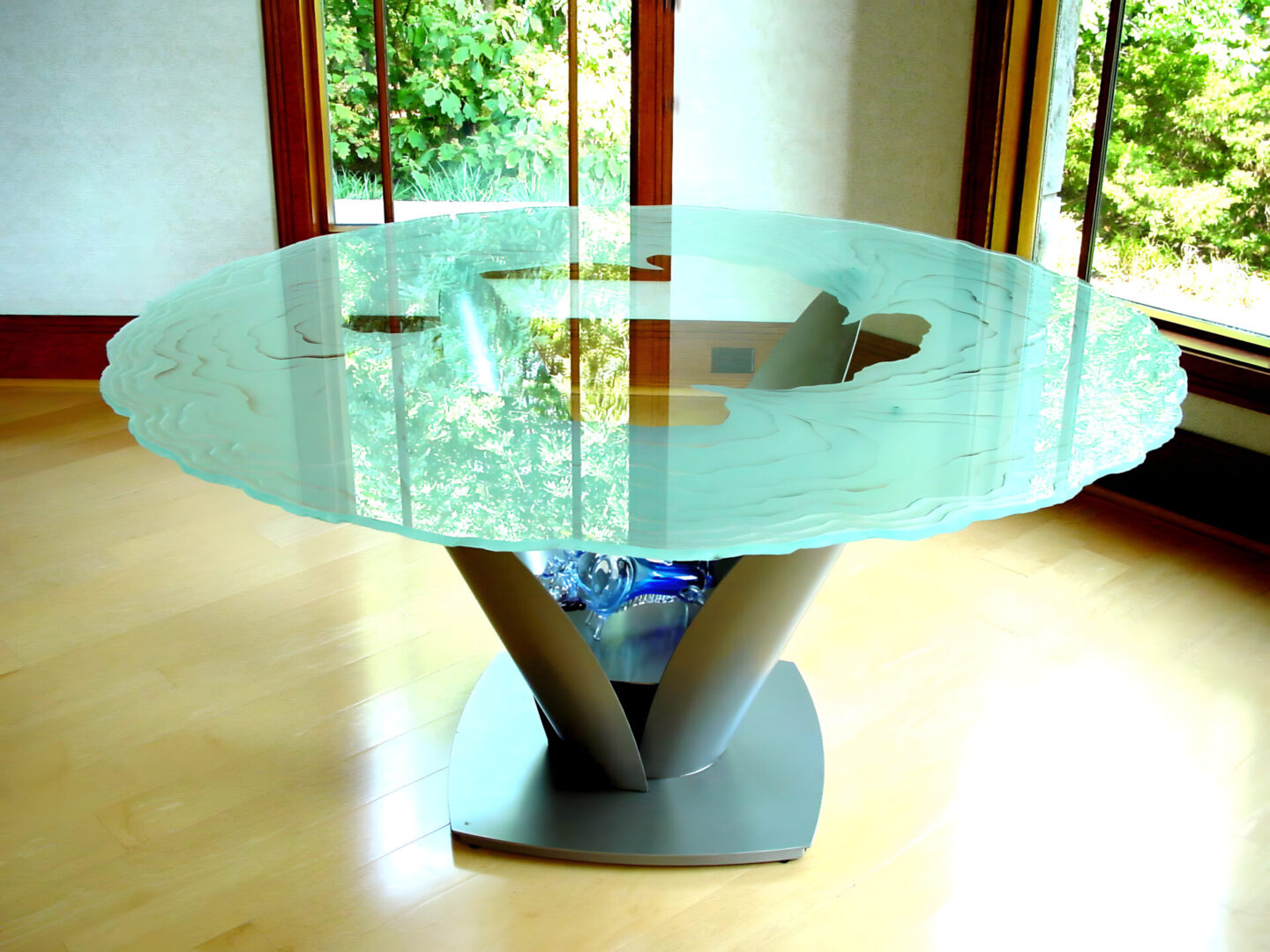 A modern glass-top table with a unique base design, placed in a room with wooden flooring and large windows.