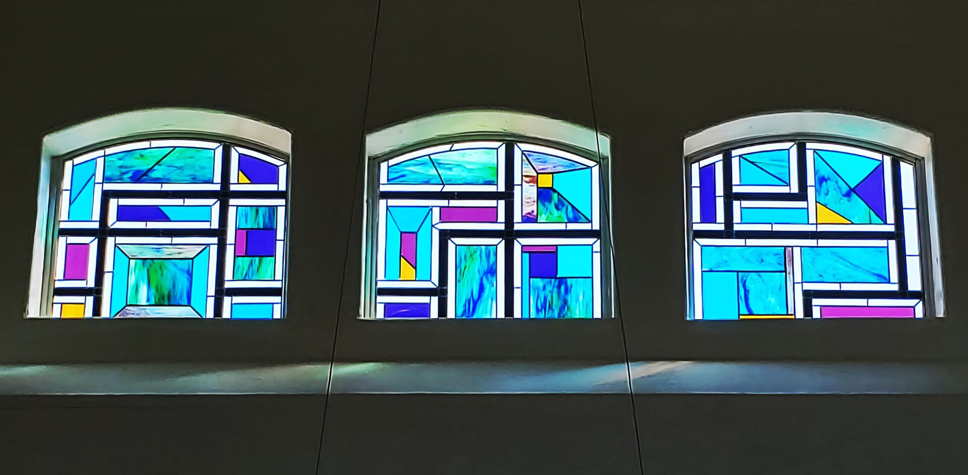 Three arched windows with colorful stained glass designs set against a gray wall.