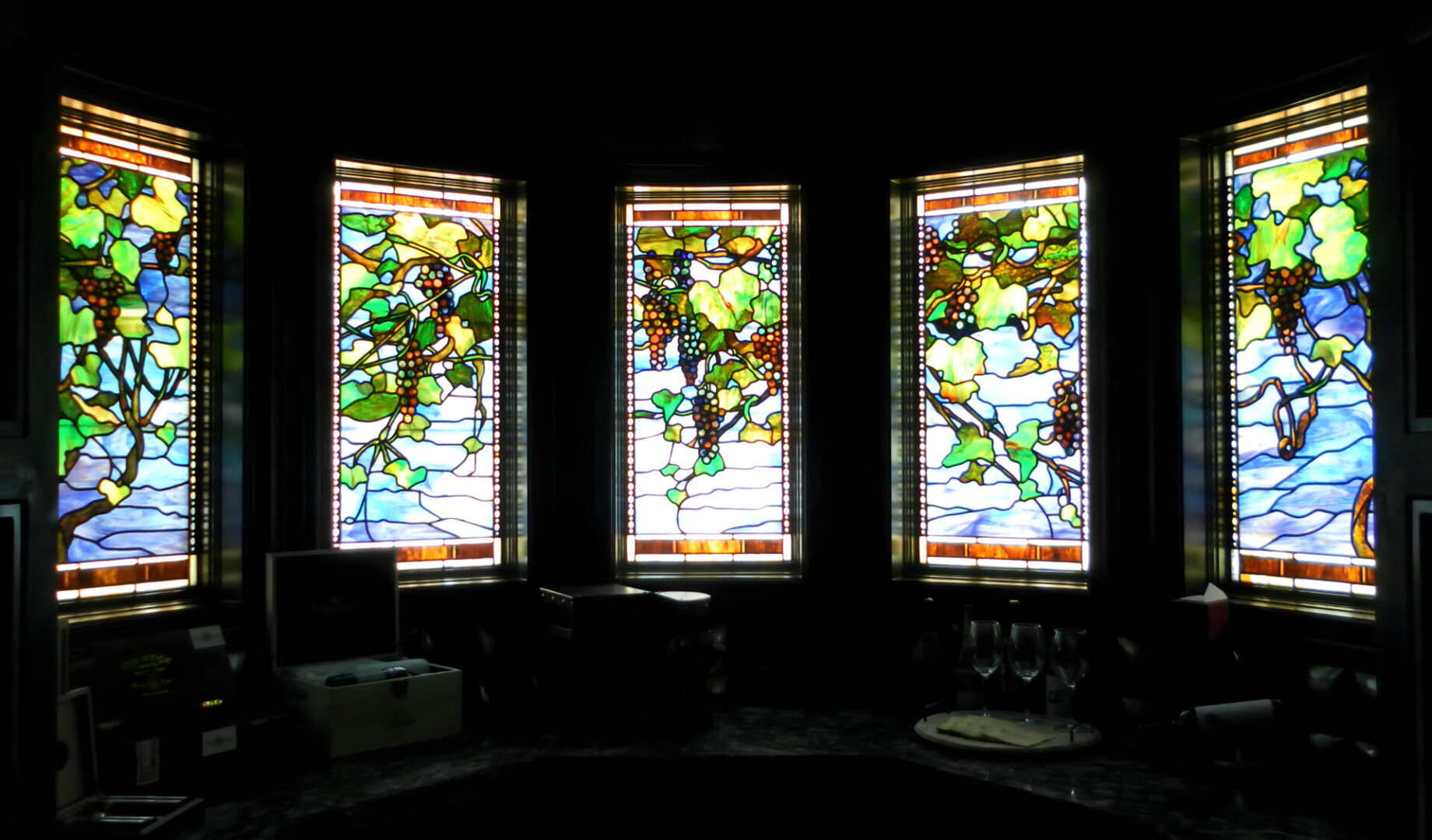 Three stained glass windows depicting trees and possibly grapevines, casting colorful light into a darkened room.