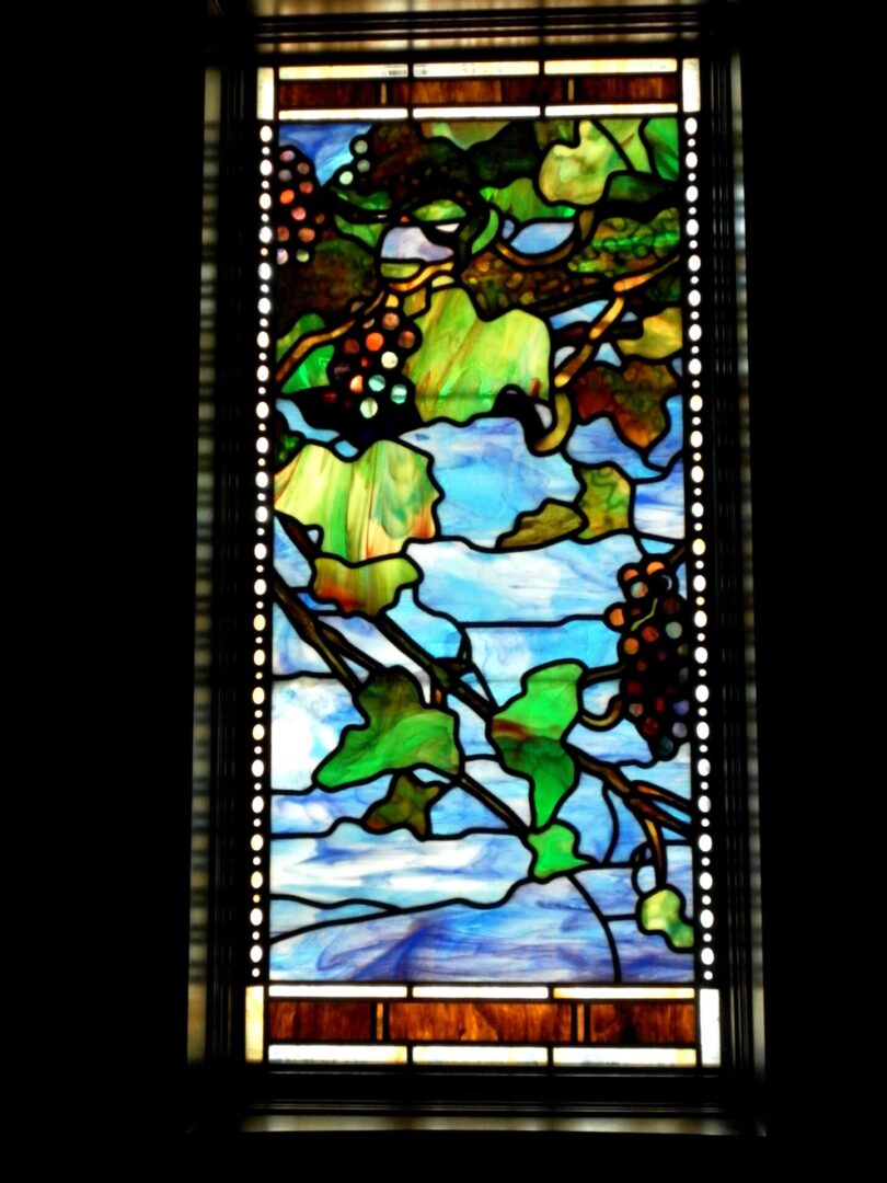 Stained glass window depicting a vine with leaves and grapes against a sky-blue background.