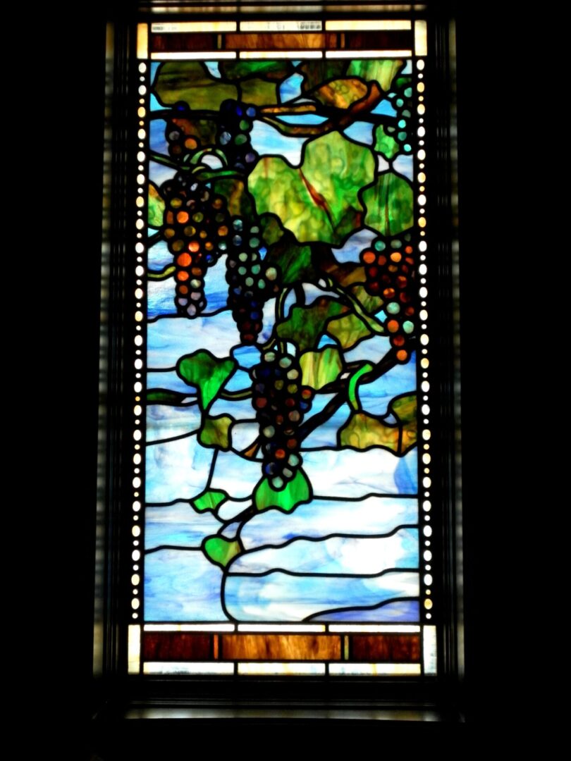 Stained glass window depicting a vine with leaves and grapes, set against a blue sky.