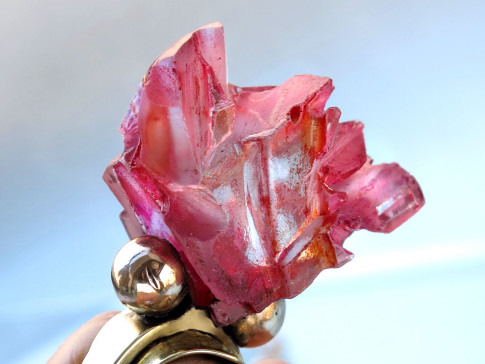 A translucent rose-pink crystal formation held by a metallic tool against a blurred background.