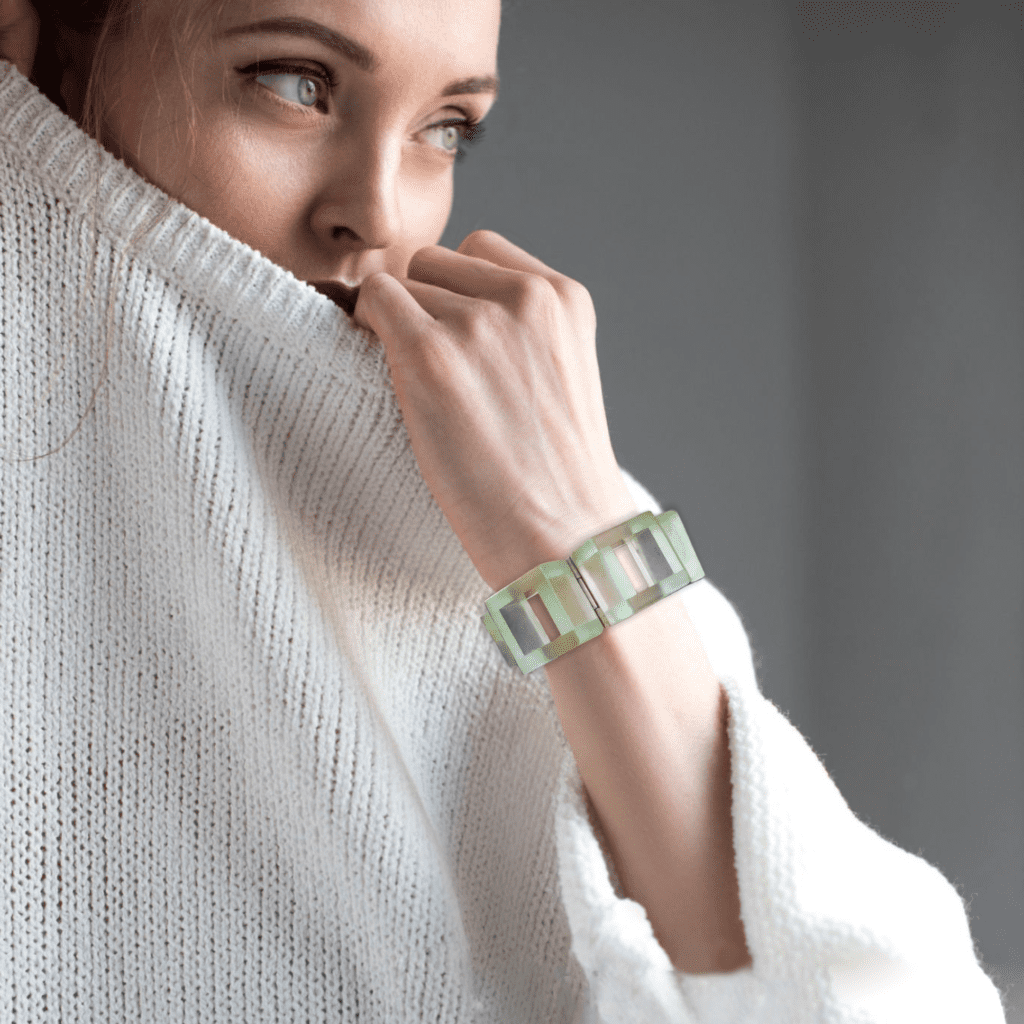 Woman in a white sweater gazing into the distance with her hand near her face, featuring a green and white bracelet.