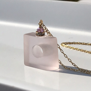 A translucent pink perfume bottle with a gold chain and a jewel-encrusted cap on a white surface.